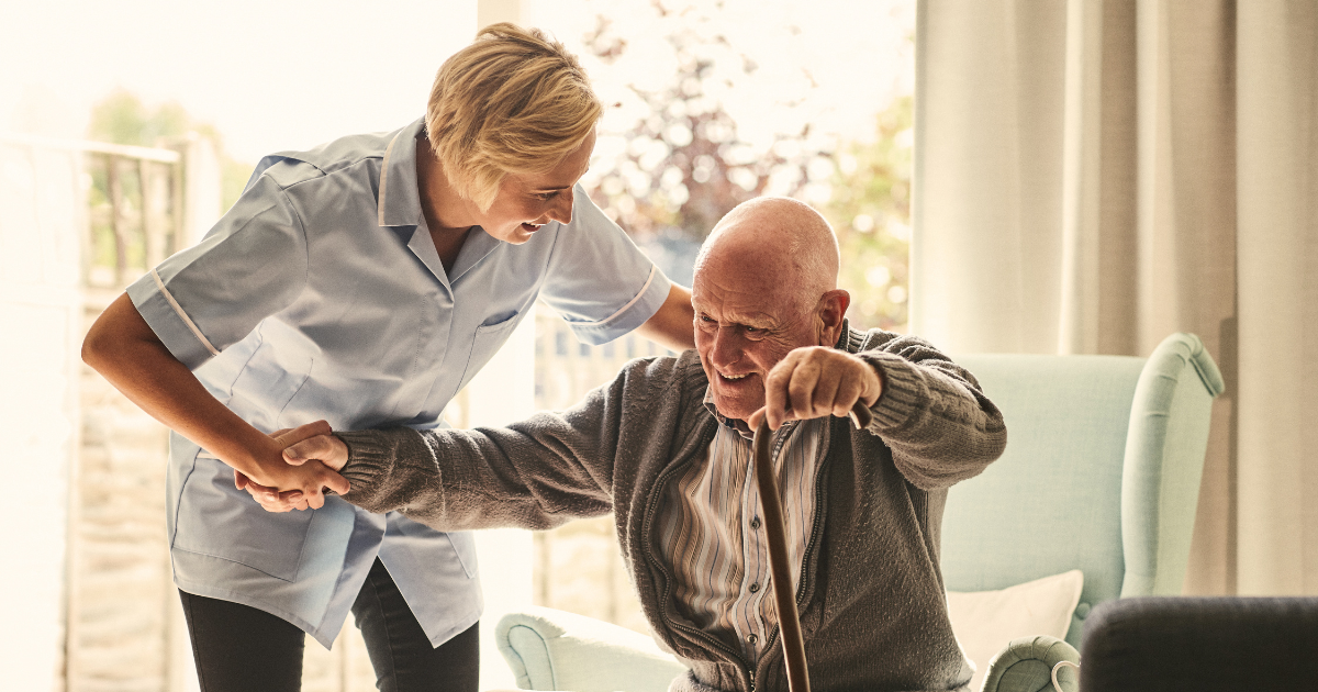 caregiver assists elderly man with cane out of his chair while providing in home care services